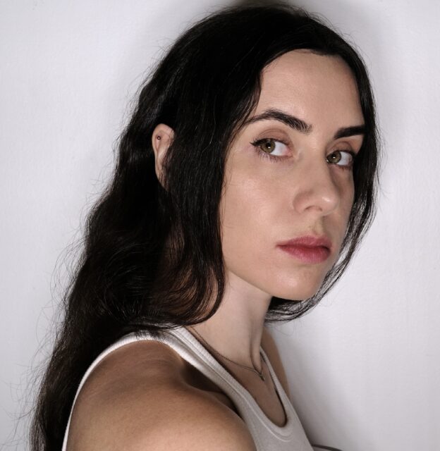 Headshot of Ira Lupu, with black hair and a white tank top, looking over her shoulder