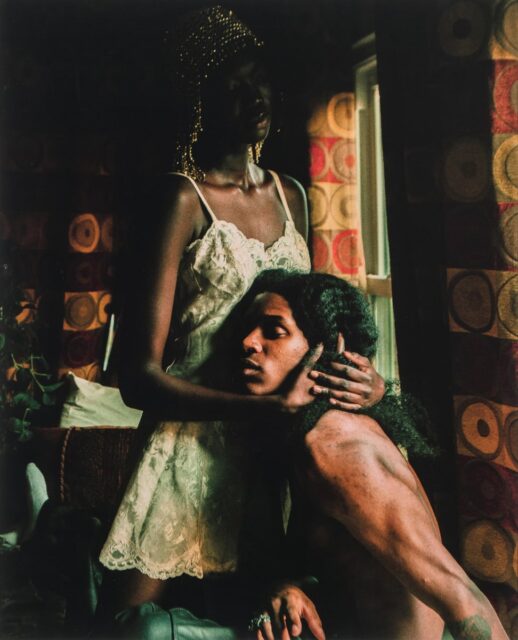 Darkly lit photograph of a Black woman in a white dress cradling a Black shirtless man