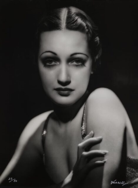 A black-and-white headshot of Dorothy Lamour a famous silver-screen actress. The photo is from the chest up and she stares directly at the viewer. Her body turned slightly away. Lamour's skin is pale with a subtle glow, her makeup is dark, and she stands out against the black background.